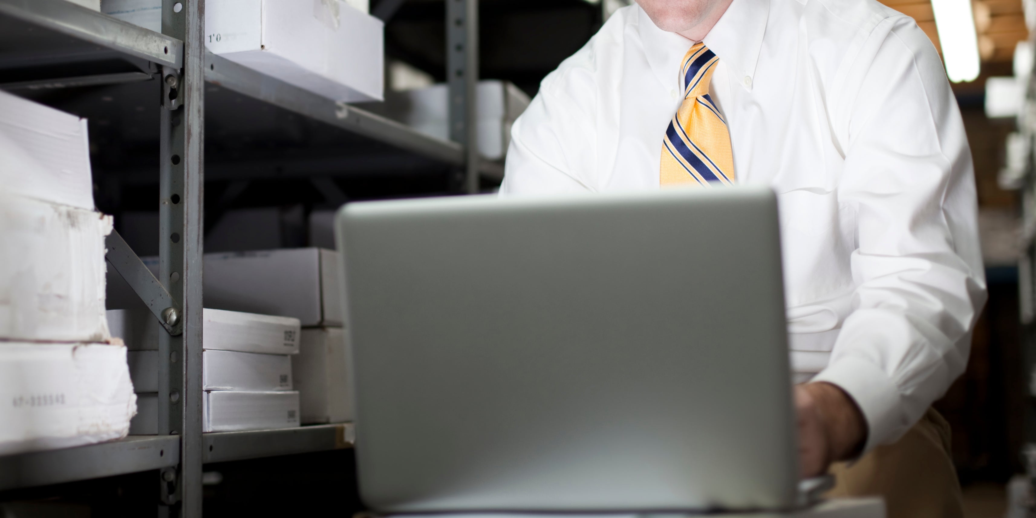 Man in a white shirt with a yellow tie works on a laptop in a storage room with shelves of boxes