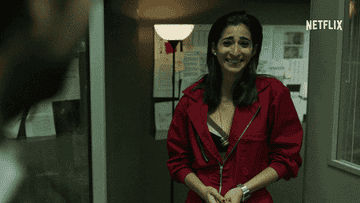Woman in red jacket smiling nervously in an office setting with bulletin boards in the background, next to the Netflix logo