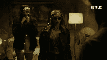 Two people in a dimly lit room wearing masks and stylish outfits, including a jacket and red sunglasses, with &quot;Netflix&quot; in the corner