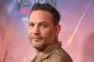 Tom Hardy with short hair and facial hair is on a red carpet wearing a patterned shirt