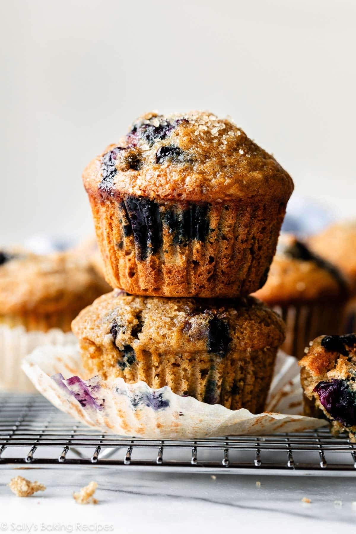 Two stacked blueberry muffins with a crumbly topping on a cooling rack, surrounded by more muffins in the background