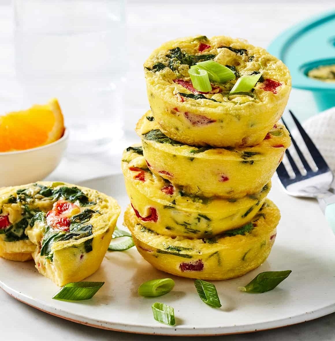 Stack of vegetable and herb egg muffins on a plate, with a glass of water and a bowl of fruit in the background