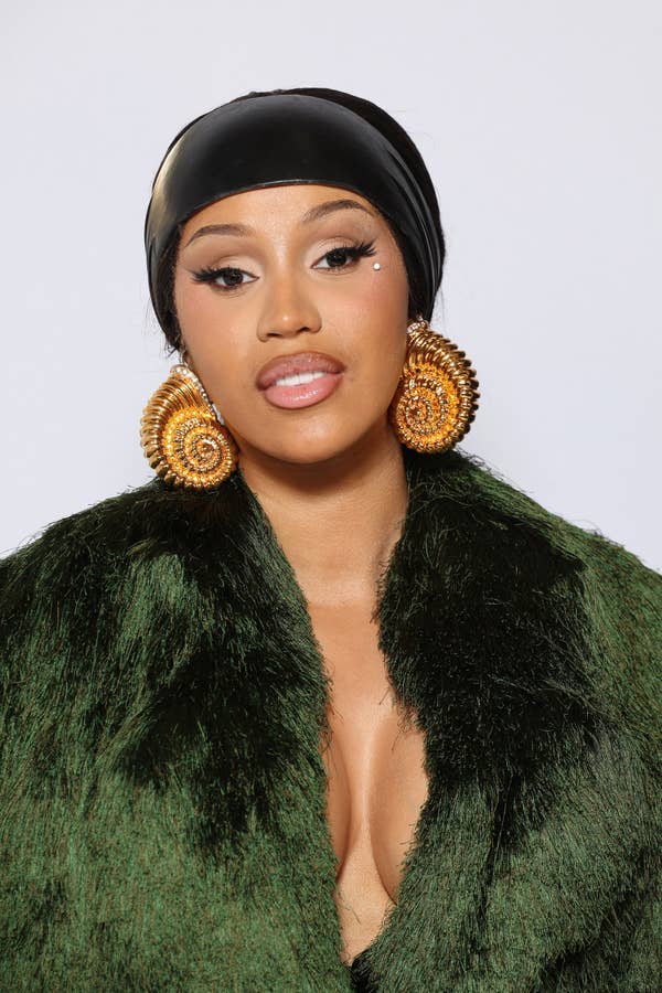 Cardi B wearing a black headscarf, large coil earrings, and a deep green faux fur coat, posing against a plain background