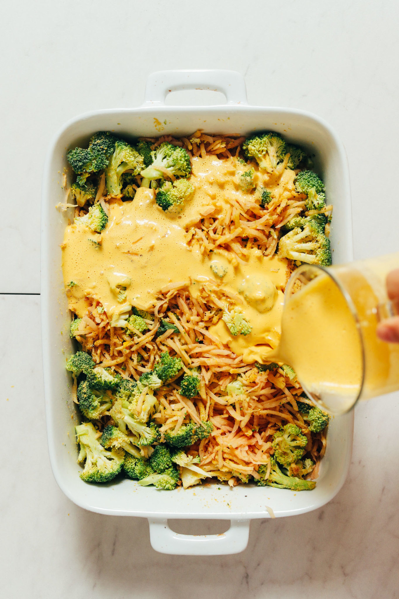 Broccoli and shredded potato casserole in a baking dish, with cheese sauce being poured on top