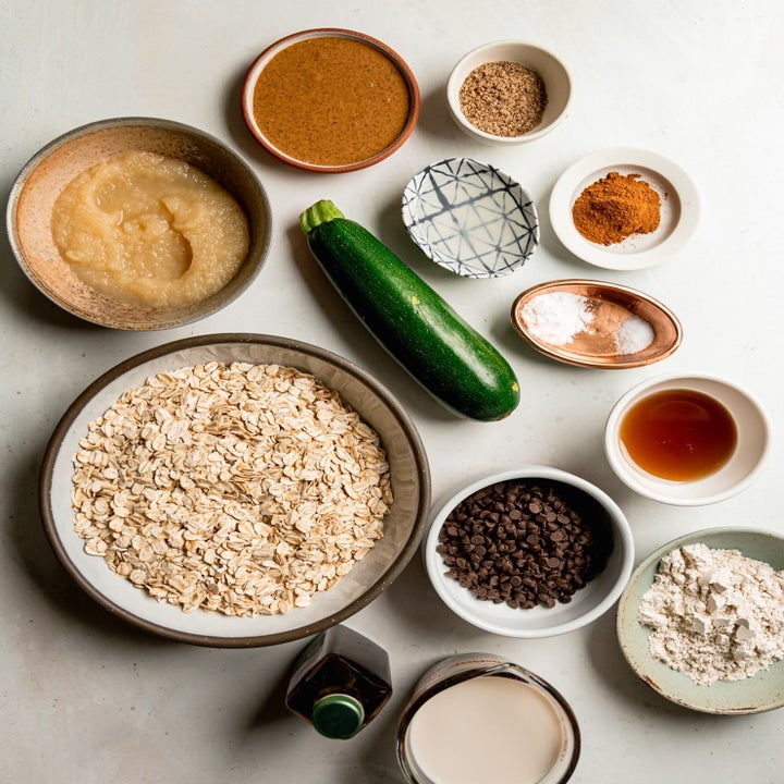Various baking ingredients including oats, zucchini, almond butter, spices, and chocolate chips arranged on a table