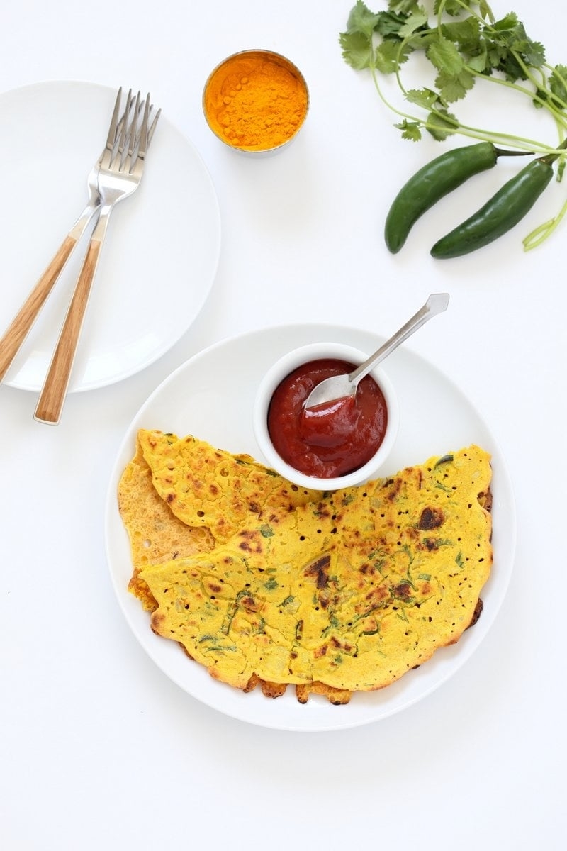 Plate with two folded savory crepes, a bowl of red sauce with a spoon, turmeric, jalapeños, and cilantro. Two forks on the side