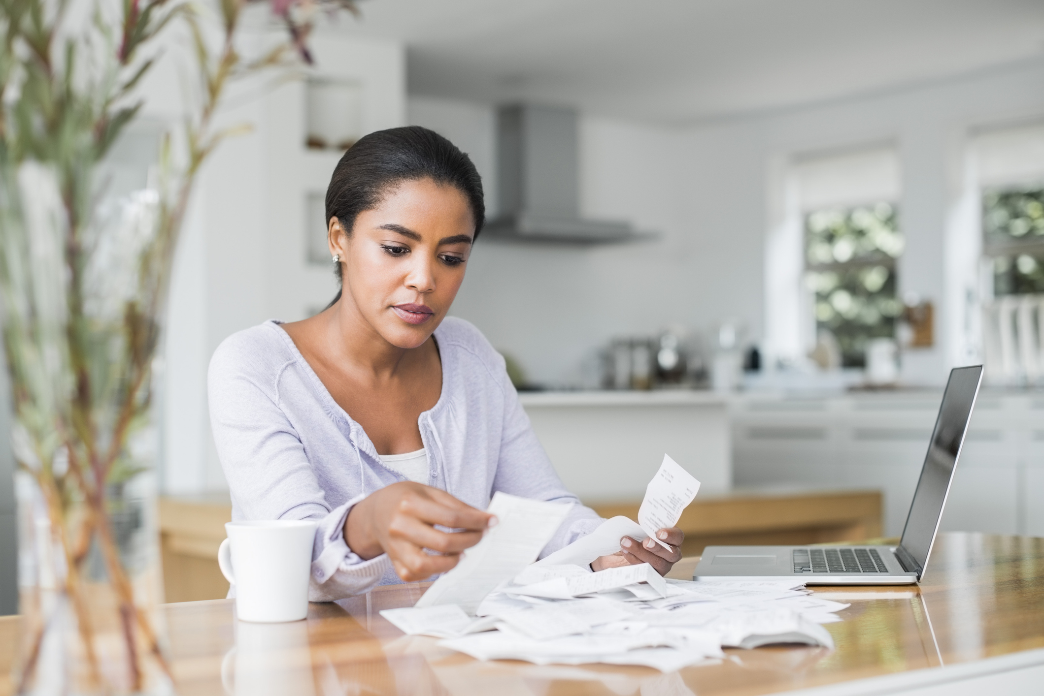 Woman reviews receipts at a kitchen table with a laptop and coffee cup nearby