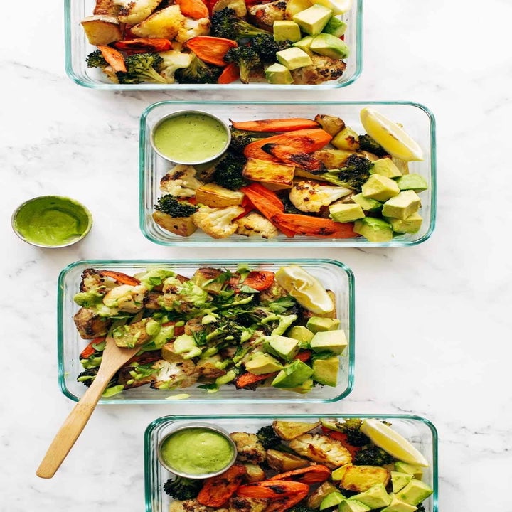 Four glass meal prep containers with roasted vegetables, avocado, and green sauce on a marble surface
