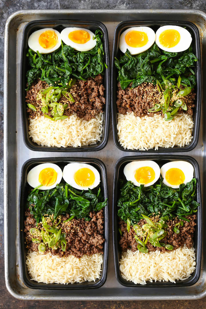 Four meal prep containers with rice, sautéed spinach, boiled eggs, ground beef, and sliced green onions on a tray