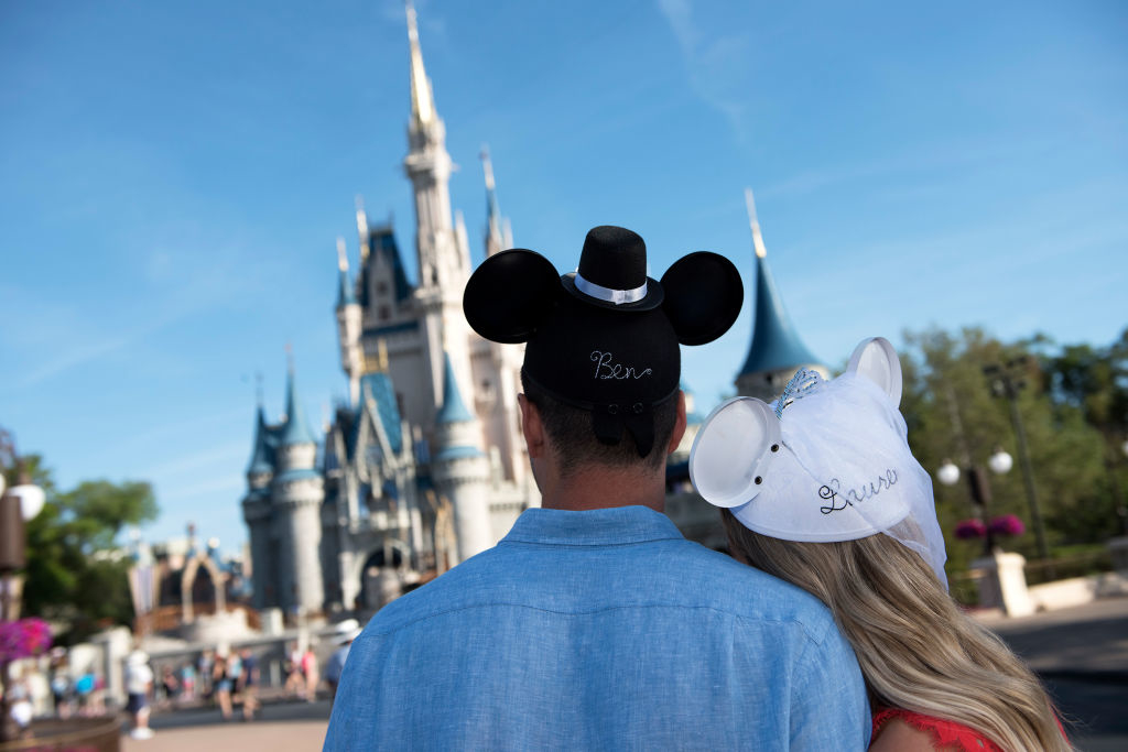Couple wearing personalized Mickey and Minnie hats, embracing in front of a castle at a theme park, symbolizing love and joy