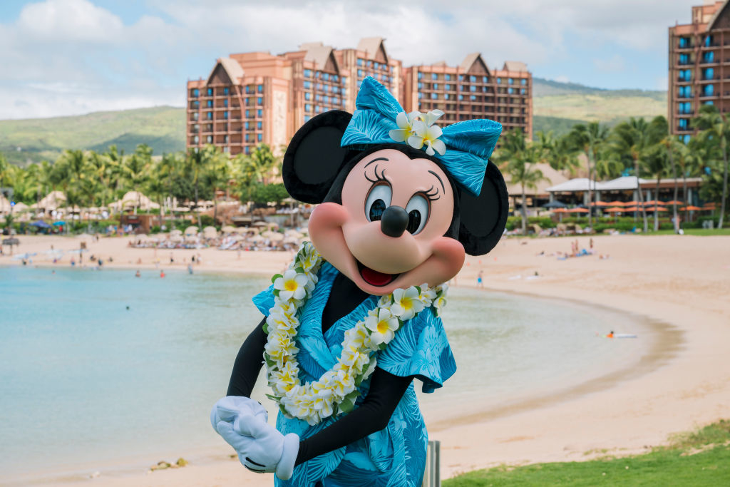 Minnie Mouse on a beach wearing a tropical-themed dress and lei, standing with resort buildings and ocean in the background