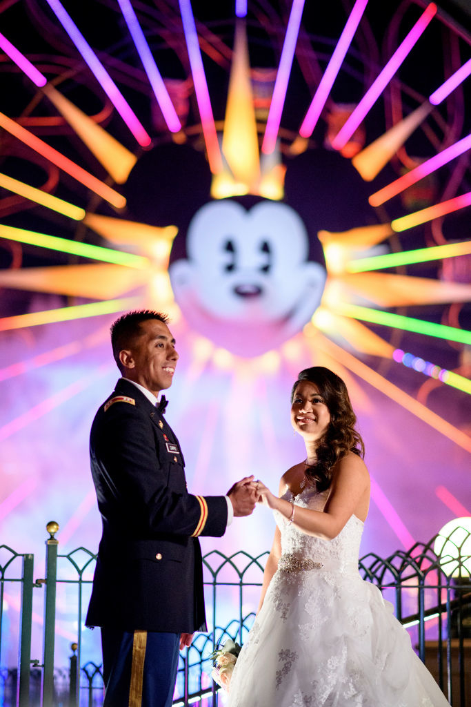 A couple in formal attire holds hands in front of a brightly lit Ferris wheel with a character's face, creating a romantic theme park backdrop