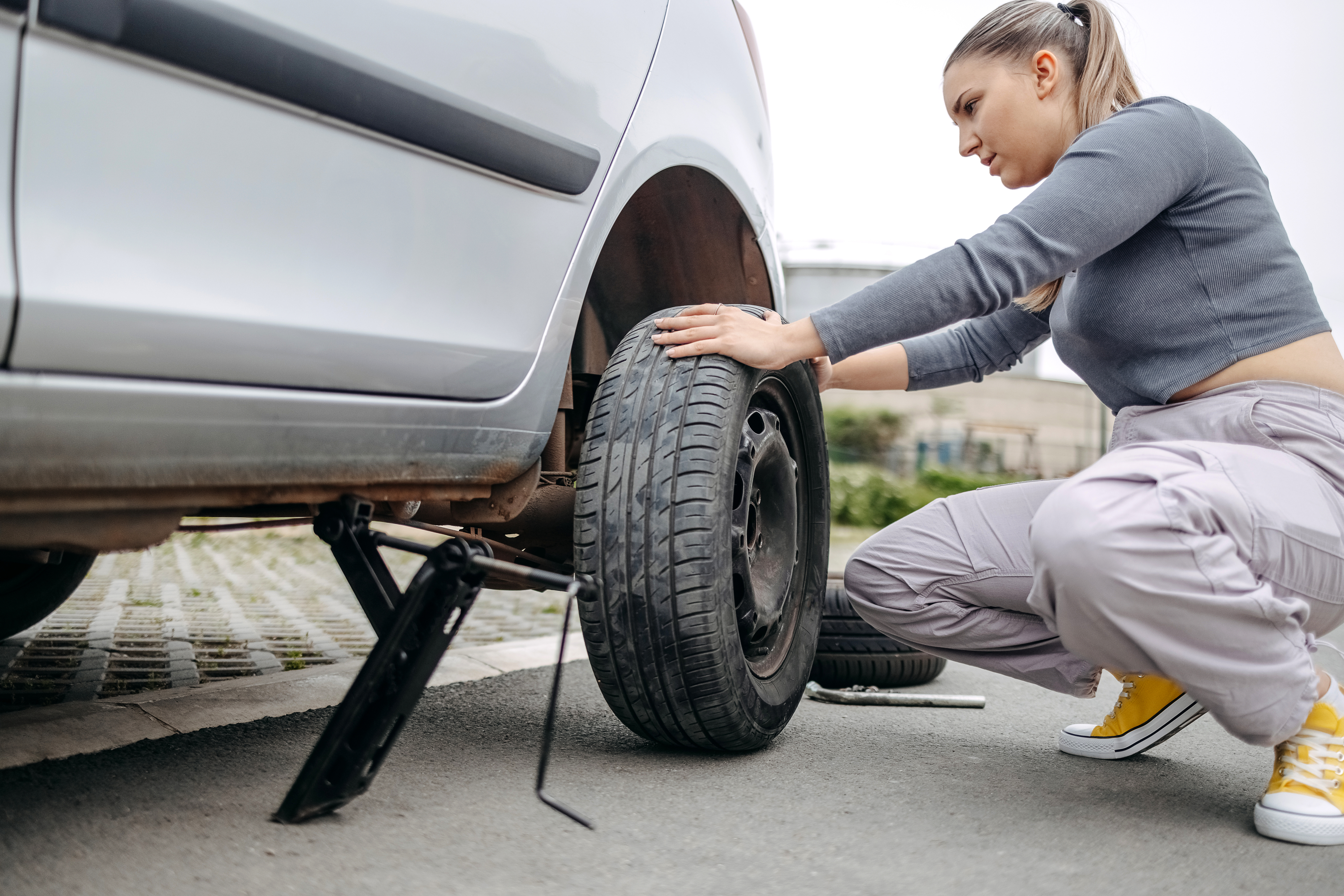 Woman kneels to change a car tire using a jack, wearing casual pants and sneakers