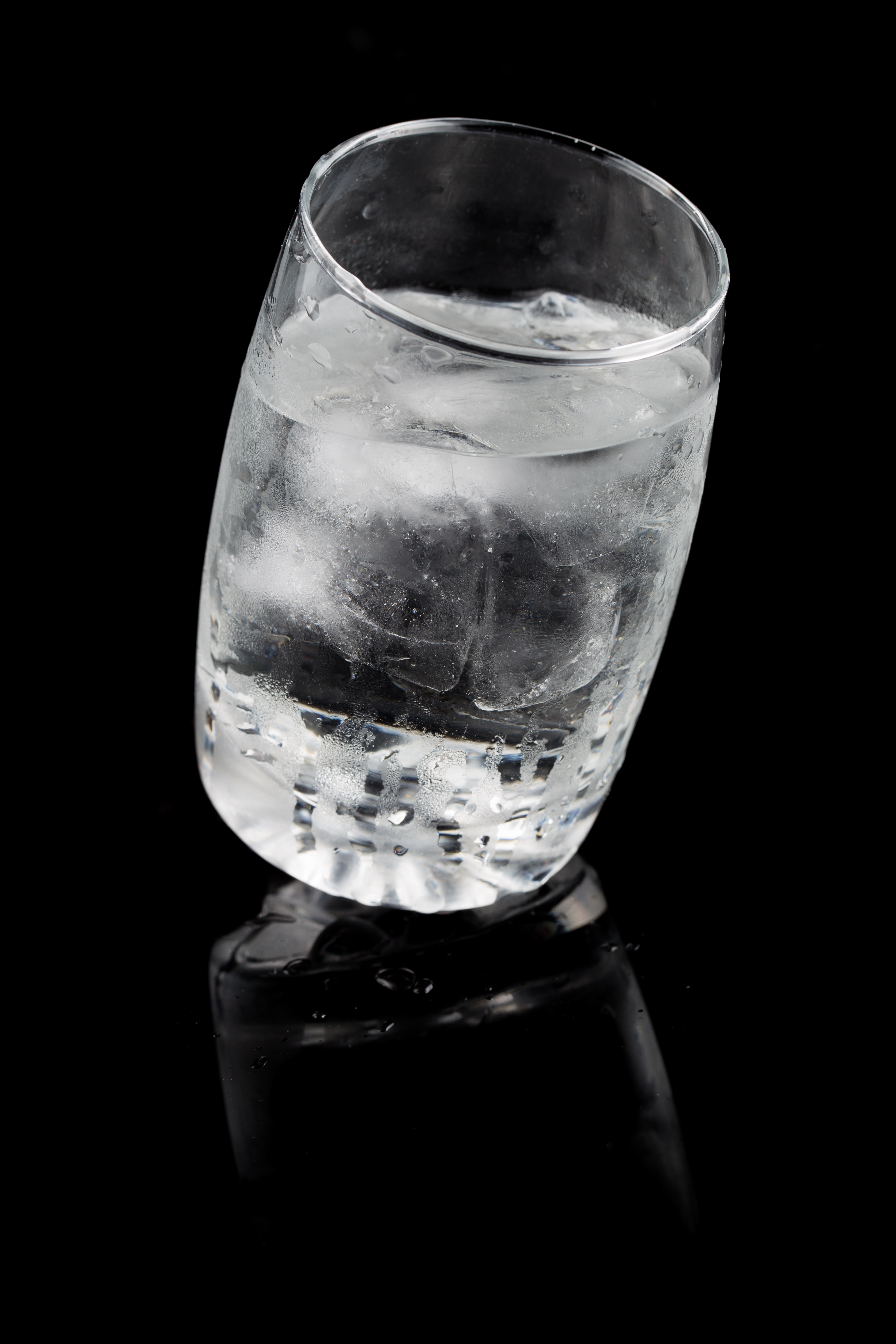 Tilted glass of ice water against a black background, reflecting on the surface