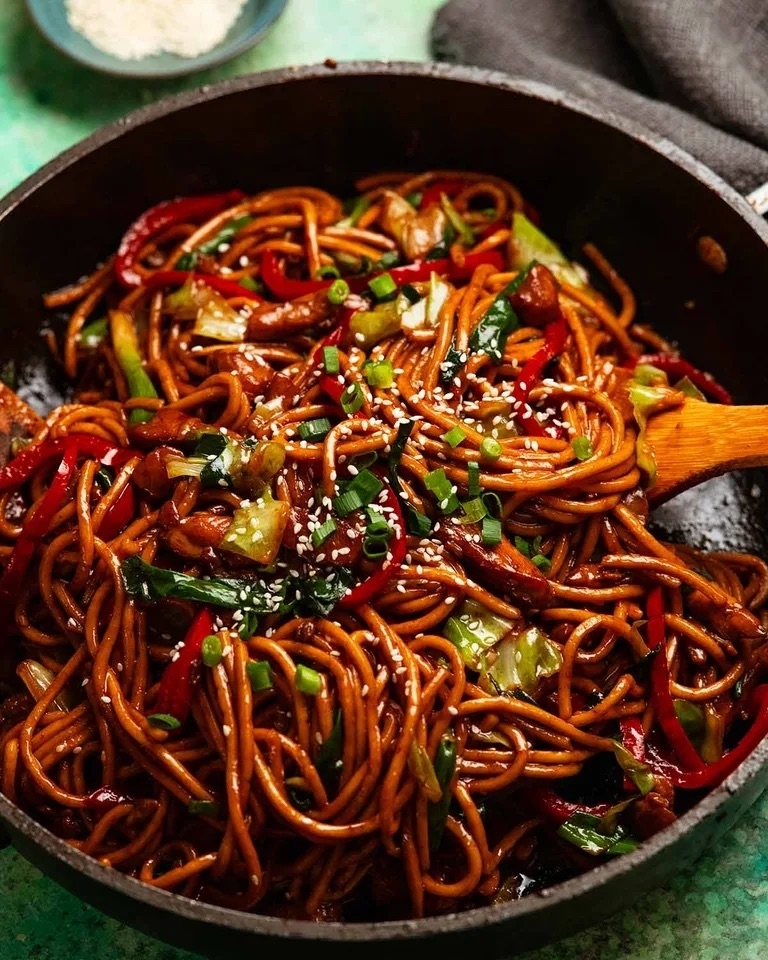 Stir-fried noodles with vegetables and sesame seeds in a pan, mixed with a wooden spoon