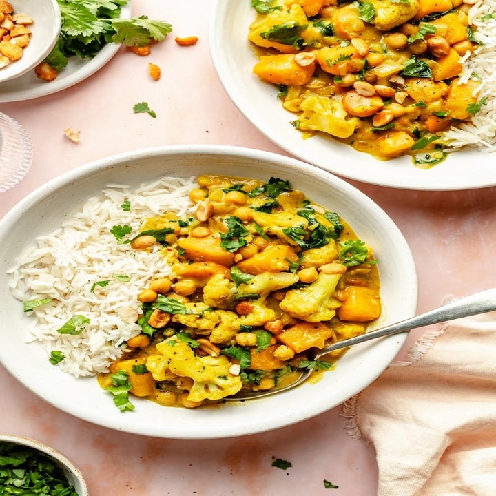 A bowl of cauliflower, chickpea, and butternut squash curry with a side of rice, garnished with cilantro and nuts