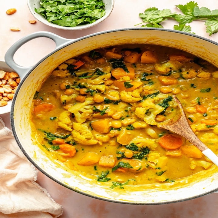 A pot of creamy vegetable curry with chickpeas and greens, with a wooden spoon, next to a bowl of fresh herbs