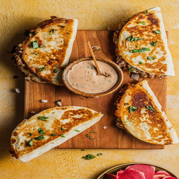 Four crispy tacos with garnishes surround a bowl of creamy dipping sauce on a wooden board