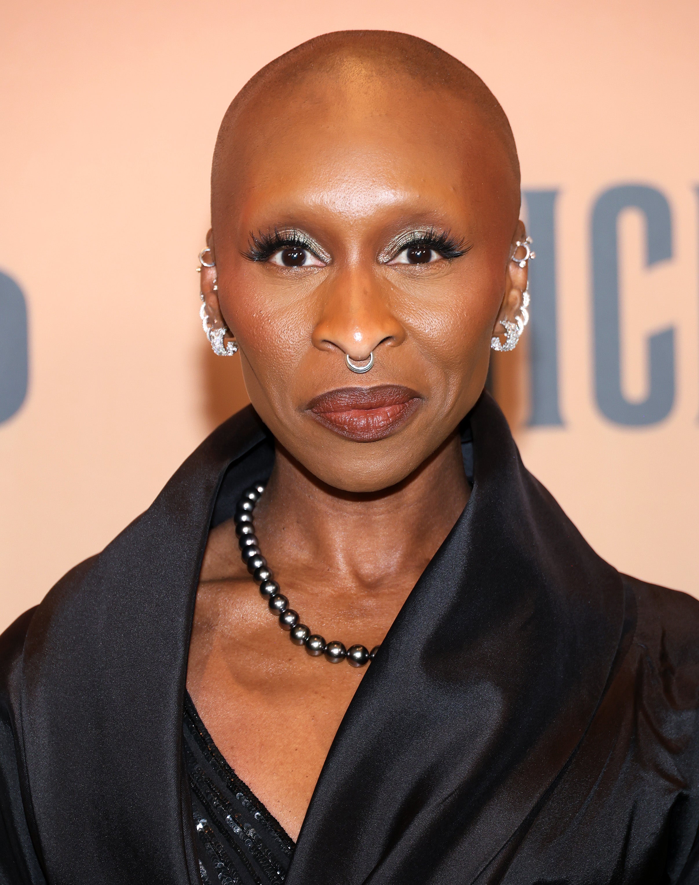 Cynthia Erivo in elegant black gown with pearl necklace, sporting multiple earrings and a nose ring, poses on a formal event backdrop