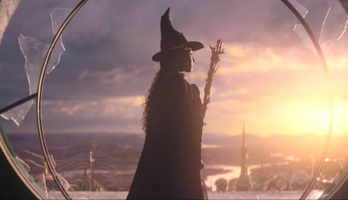 Cynthia Erivo figure in a wizard hat holds a staff, gazing at a dramatic sunset through a circular window.