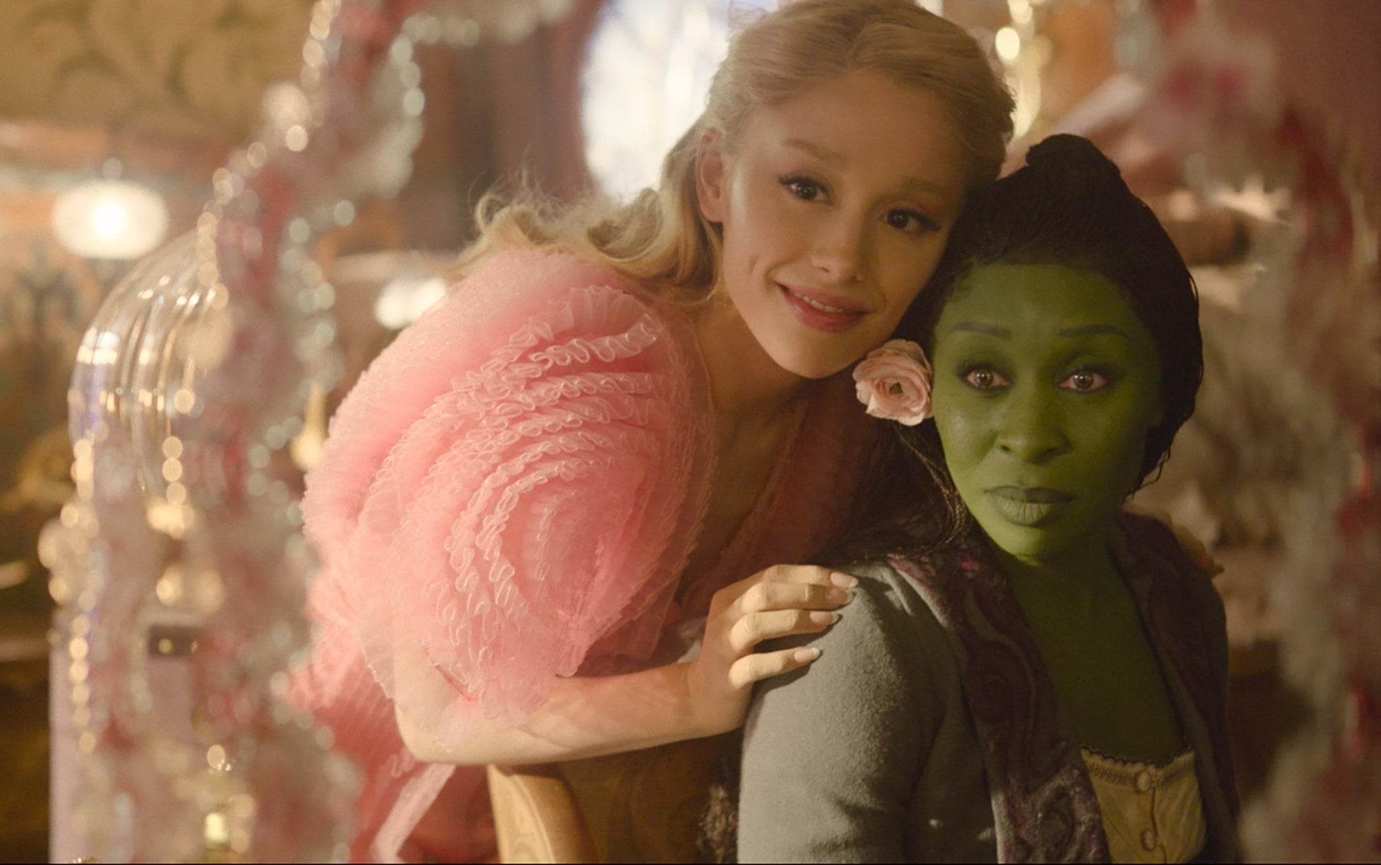 Two people in an elegant room; one with light hair wearing a fluffy top, and the other with green skin and a flower in their hair