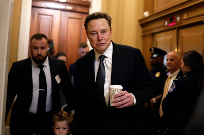 Elon Musk in a suit holding a coffee cup walks in a crowded hallway with security and onlookers