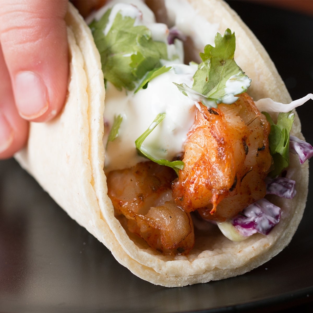 Close-up of a shrimp taco with cilantro, cabbage, and a creamy sauce in a soft tortilla, held in a hand