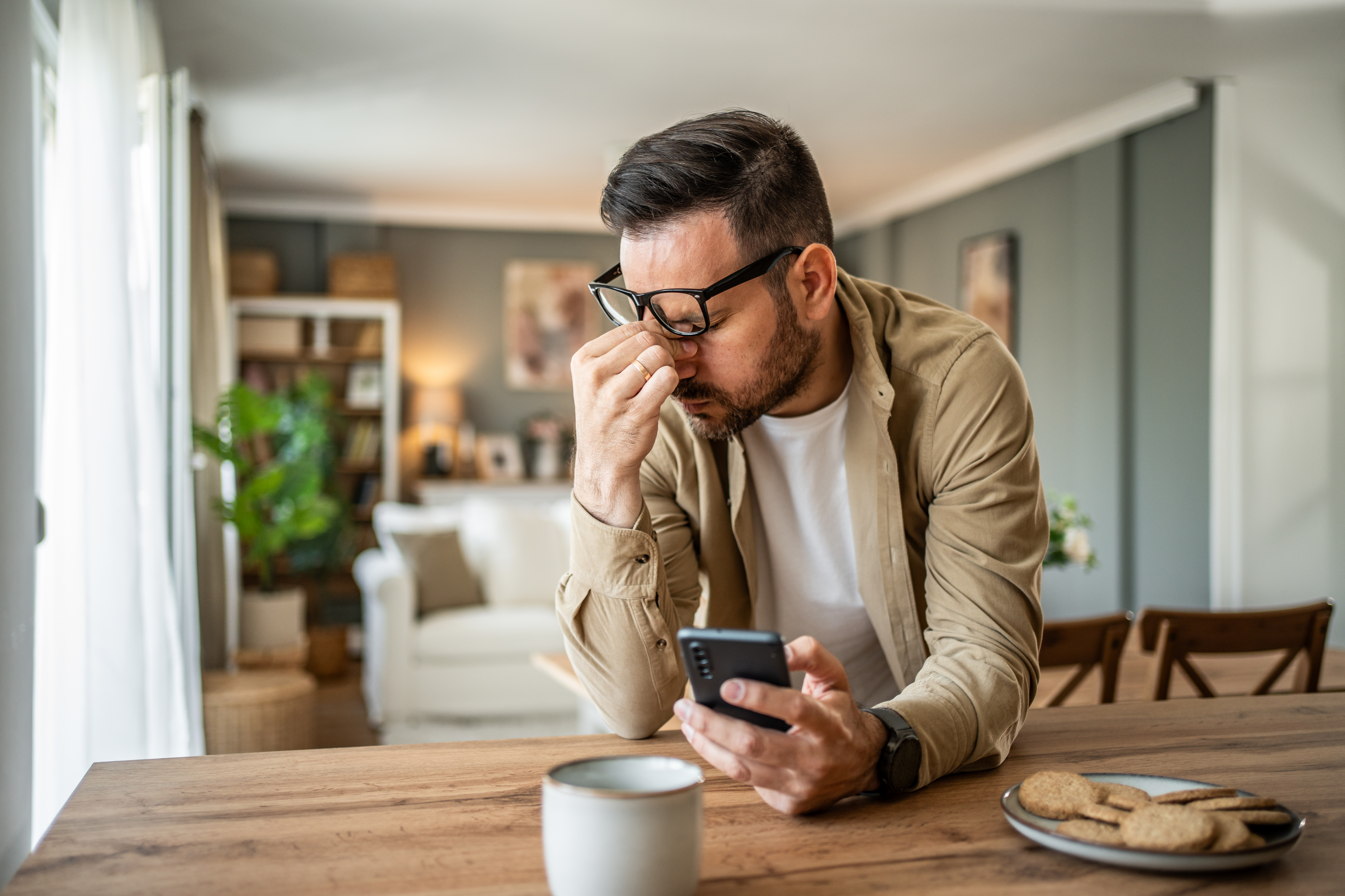 Man in casual attire looks stressed while holding a smartphone at a table, with a plate of cookies beside him in a living room