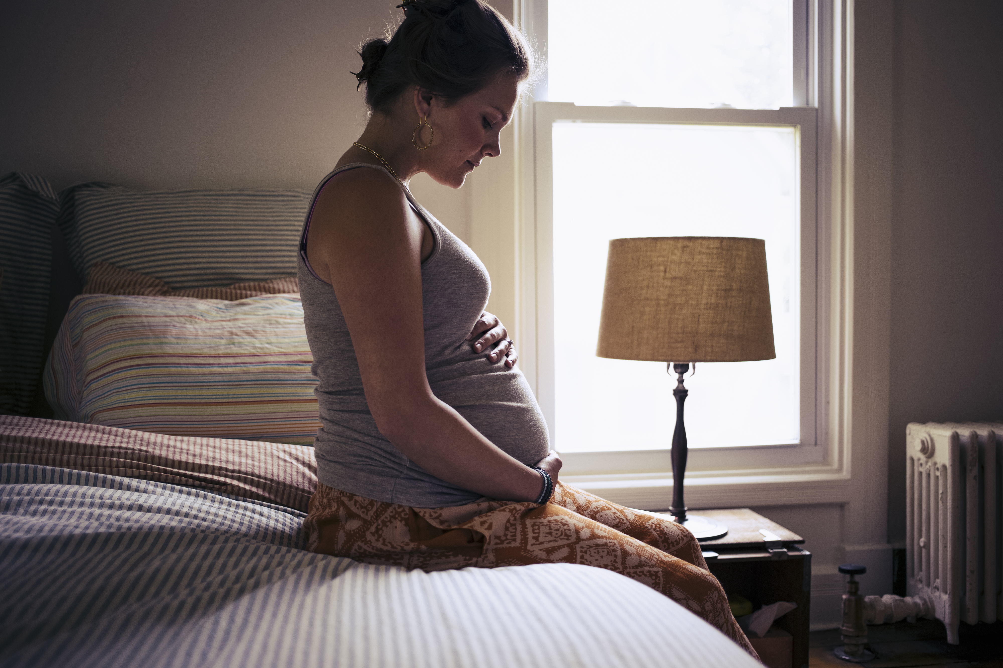 A pregnant woman sits on a bed, gently holding her belly, in a softly lit bedroom