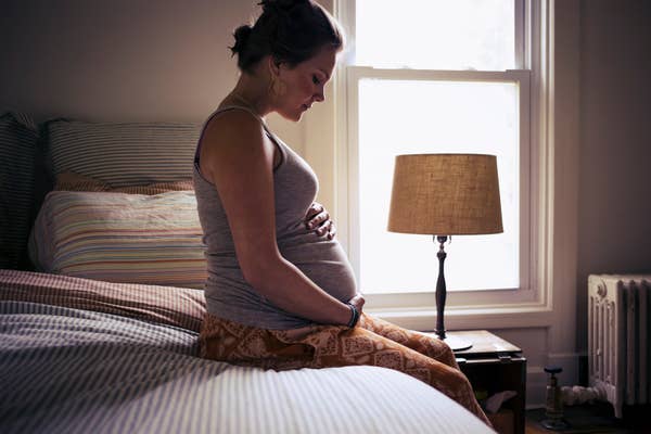 A pregnant woman sits on a bed, gently holding her belly, in a softly lit bedroom