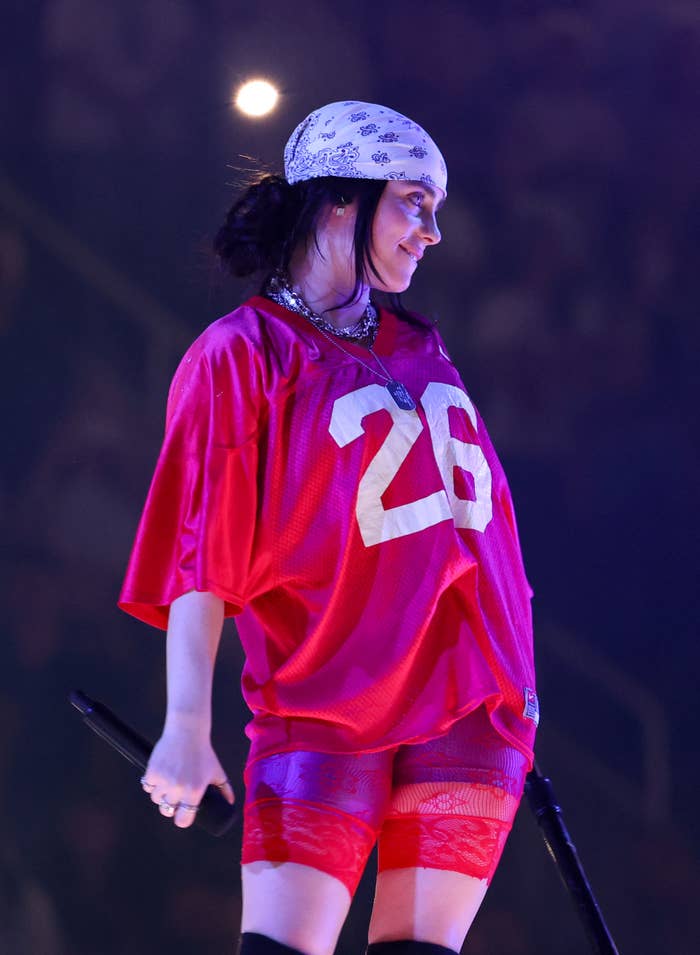 Person on stage wearing a loose sports jersey with the number 26, lace shorts, and a patterned headscarf, holding a microphone