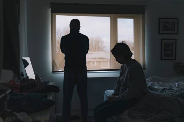 Two people in a dimly lit bedroom, one standing by a window and the other sitting on a bed, both in contemplative poses