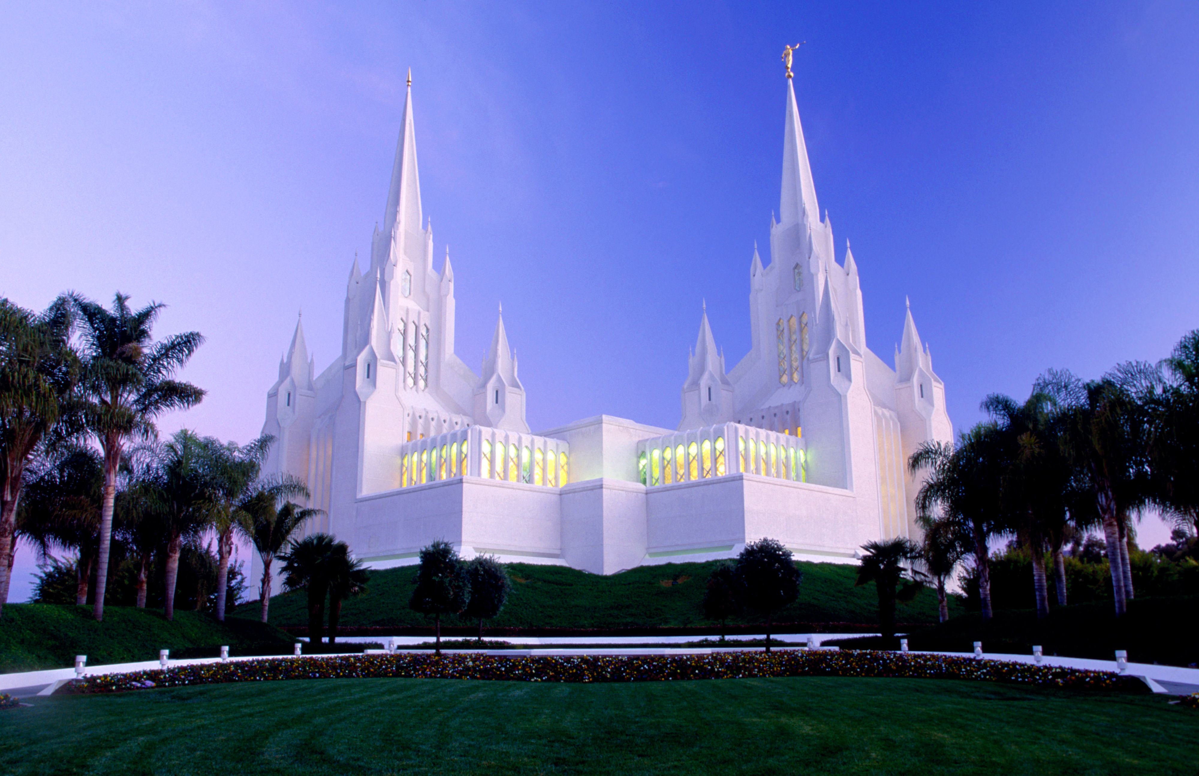 White Gothic-style temple with tall spires against a clear sky, surrounded by palm trees and green lawns