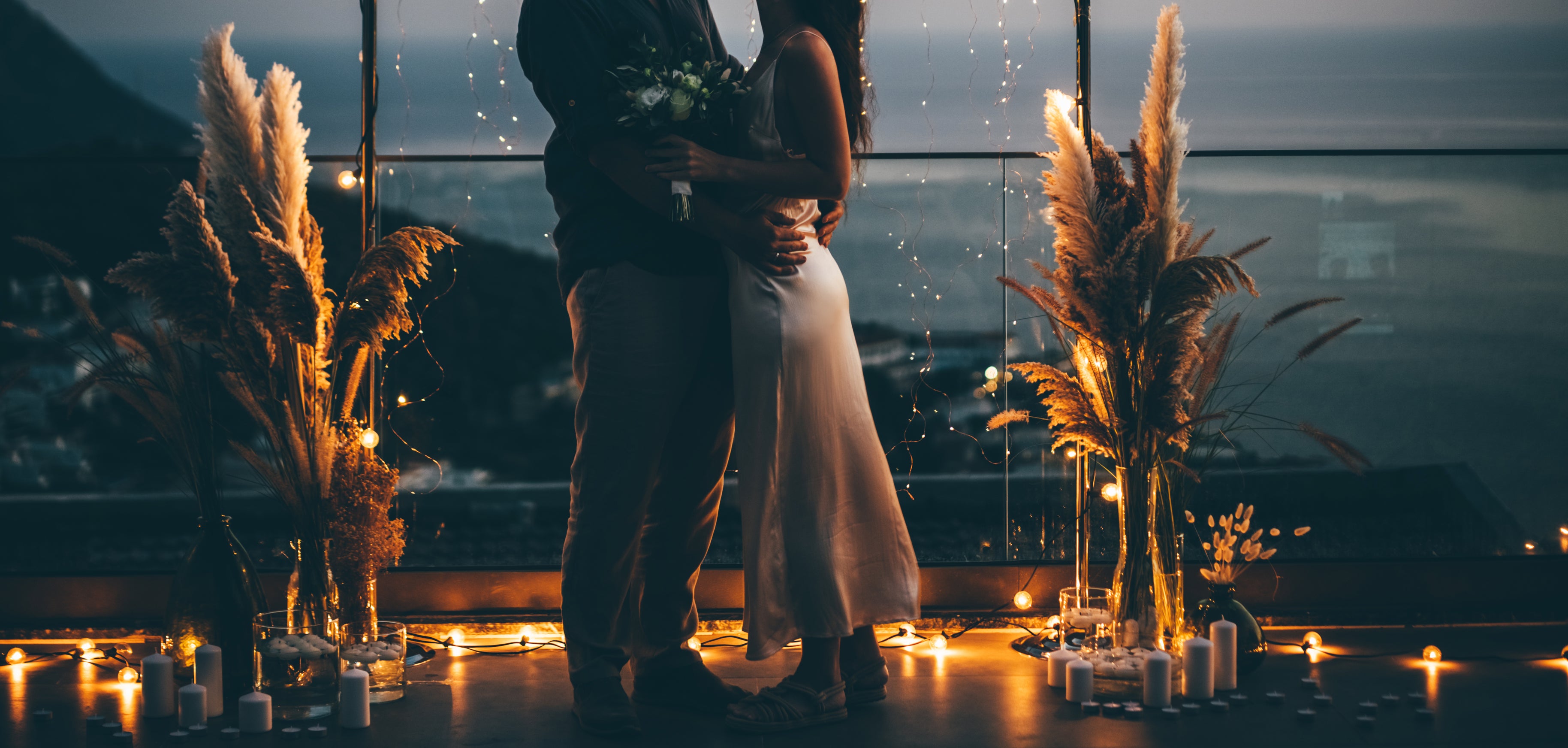 A couple embraces on a balcony at sunset, surrounded by string lights and pampas grass decorations. Romantic ambiance with candles