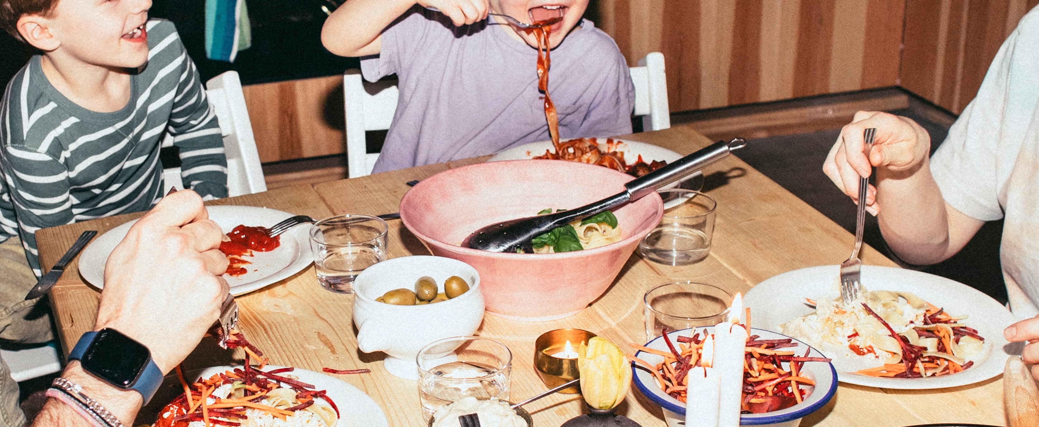 Family enjoying dinner, two children playfully eating pasta, smiling with an adult at a cozy table. Laughter and joy fill the room