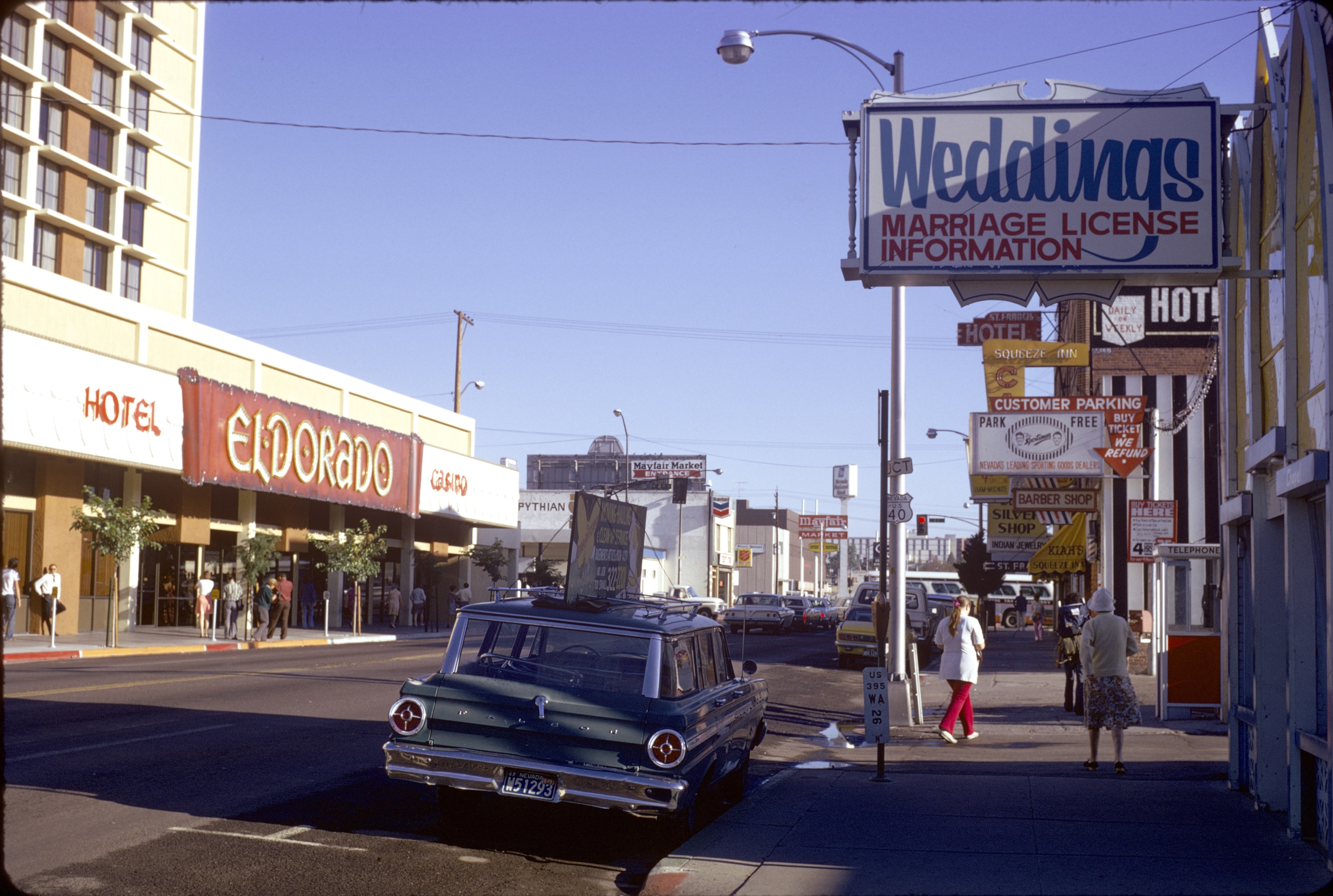 Busy city street with vintage car and storefronts, including Eldorado Hotel and a wedding license information sign. People walking on the sidewalk