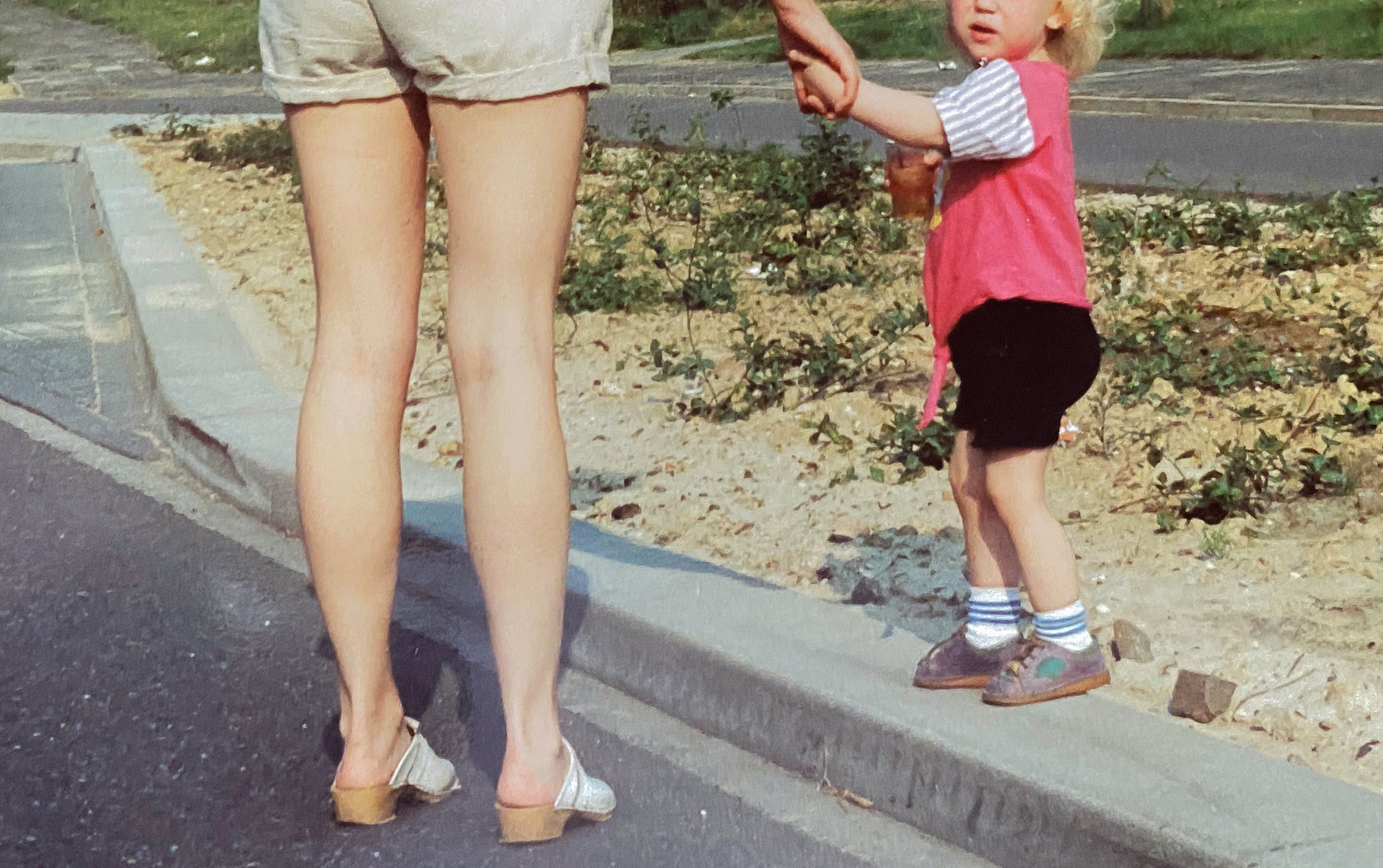 Adult holding a child's hand while walking on a roadside path, surrounded by greenery
