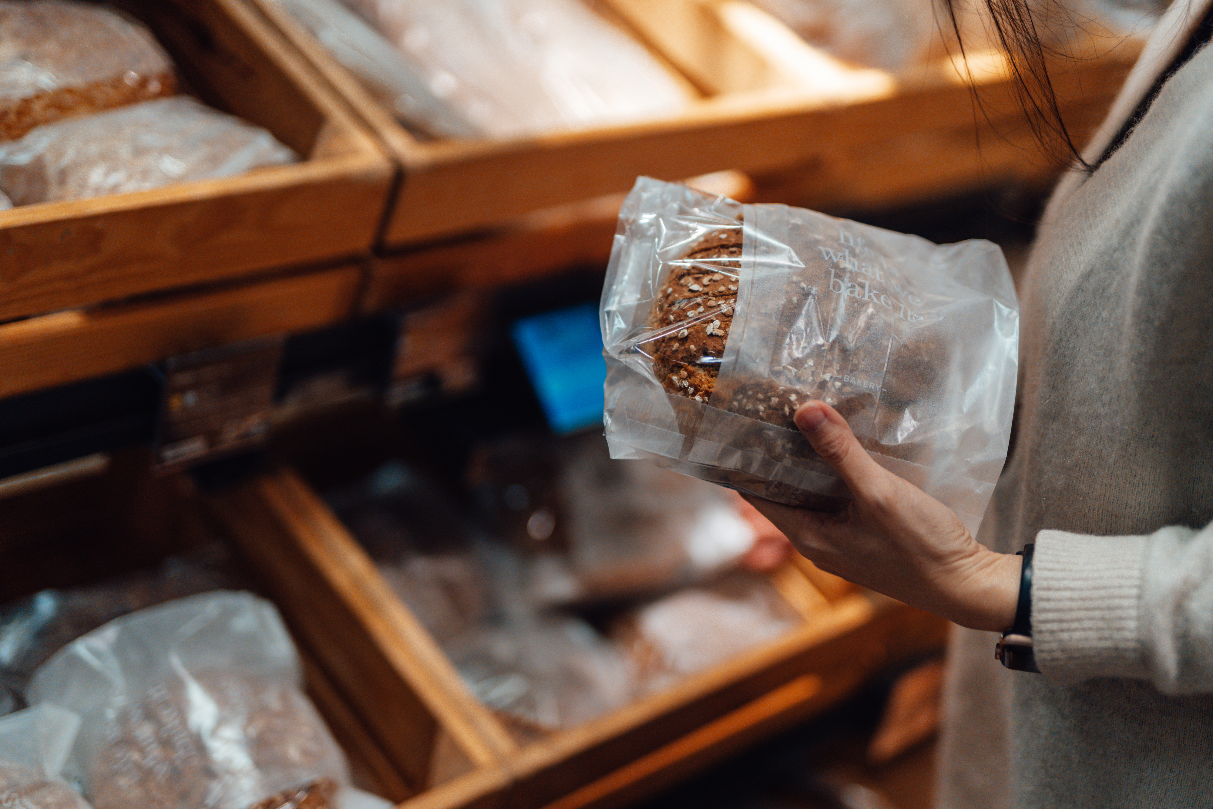 Person holding packaged bread loaf in a bakery aisle, surrounded by shelves of bread