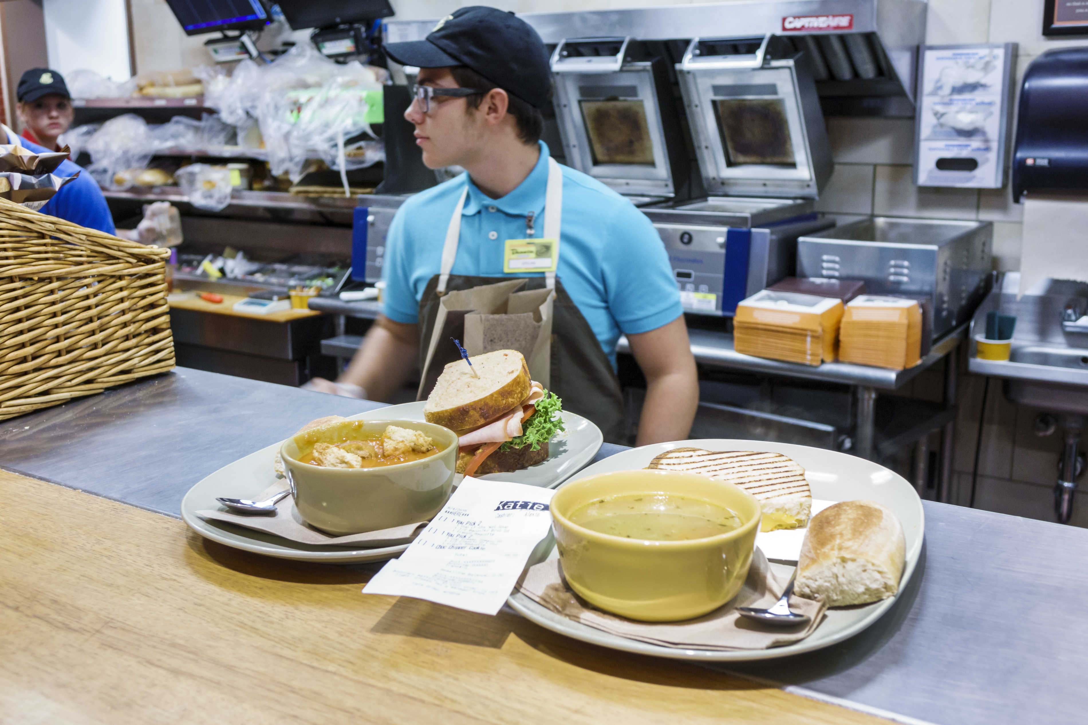 Café counter with a staff member serving sandwiches and soups on trays, ready for customers