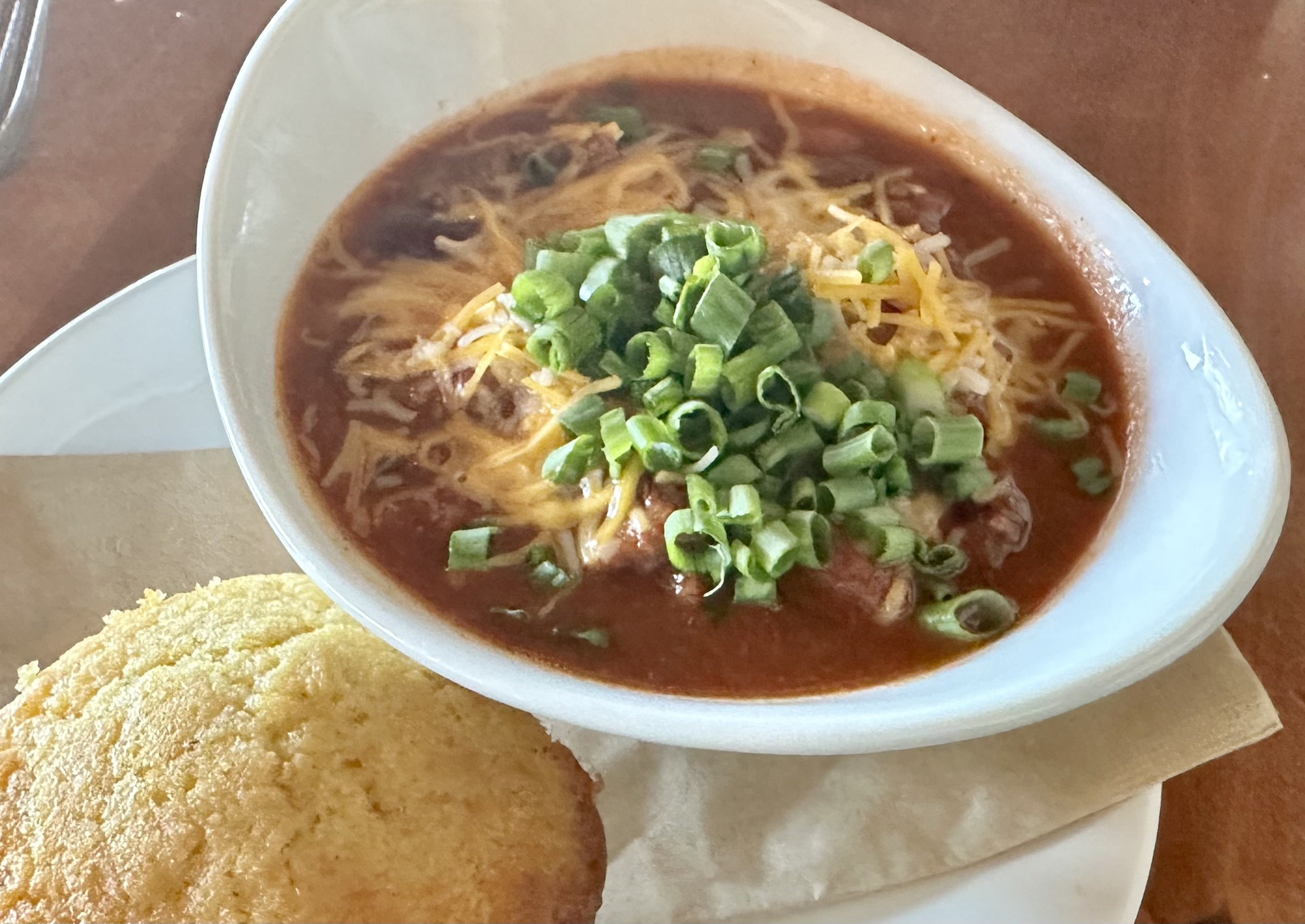 Bowl of chili topped with shredded cheese and diced green onions, served with a side of cornbread on a napkin