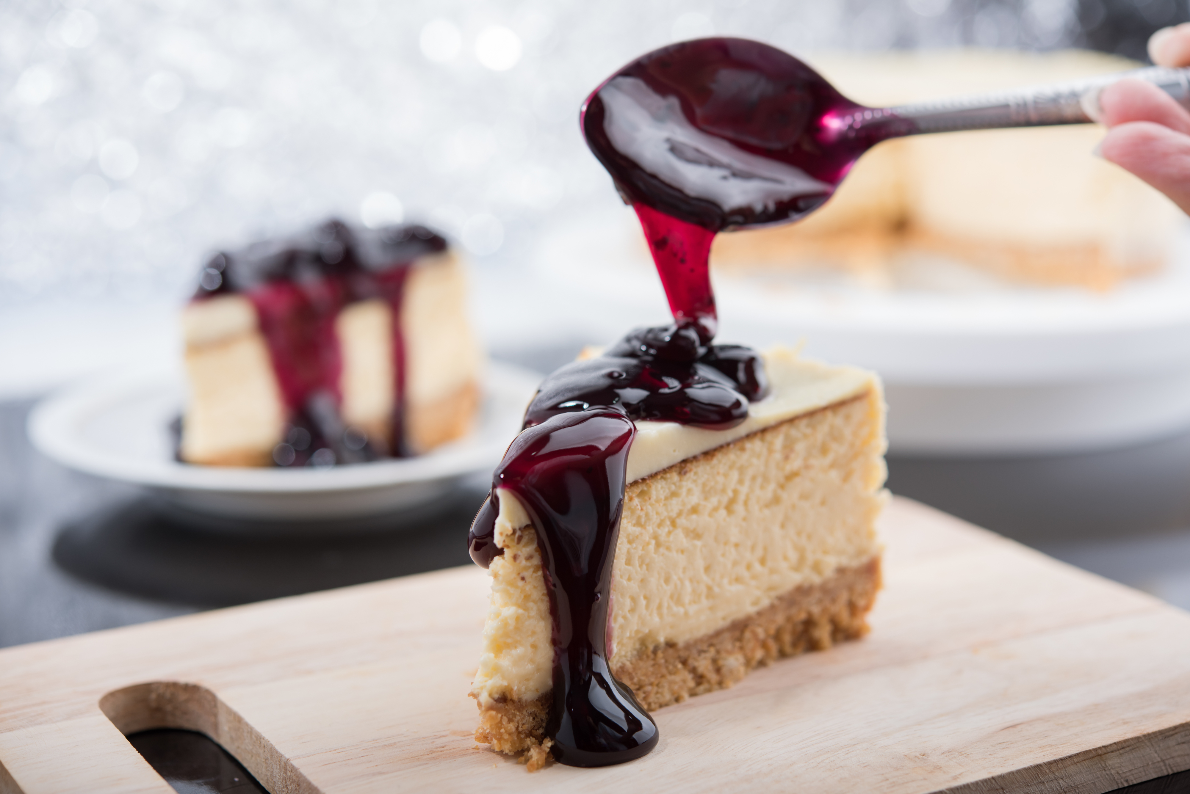 Cheesecake slice on a wooden board, topped with a berry sauce being spooned over it, with another slice on a plate in the background