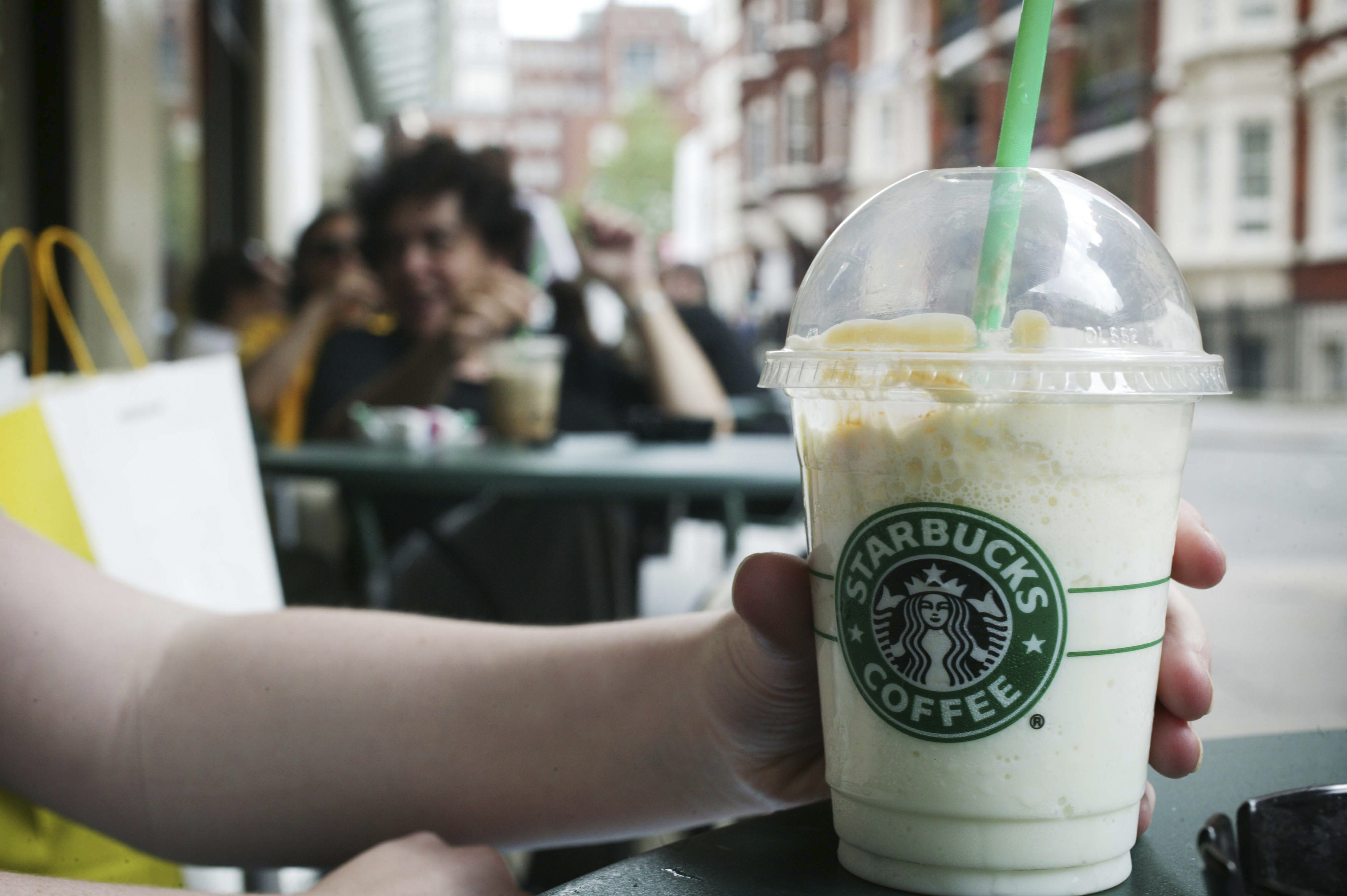 A person holds a Starbucks cup outside at a café table, with people and buildings in the background