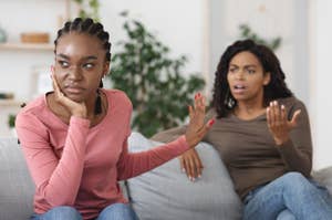 Two women sitting on a couch; one looks frustrated with her hand up, the other gestures as if explaining. They seem to be having a disagreement