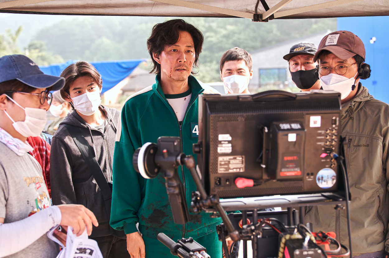 Actors and crew, some wearing masks, on a film set; a man in a numbered tracksuit stands in front of a monitor