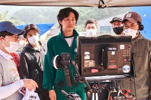 Actors and crew, some wearing masks, on a film set; a man in a numbered tracksuit stands in front of a monitor