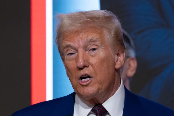 男性がスーツを着て話している。背景にも別の人物がいる。