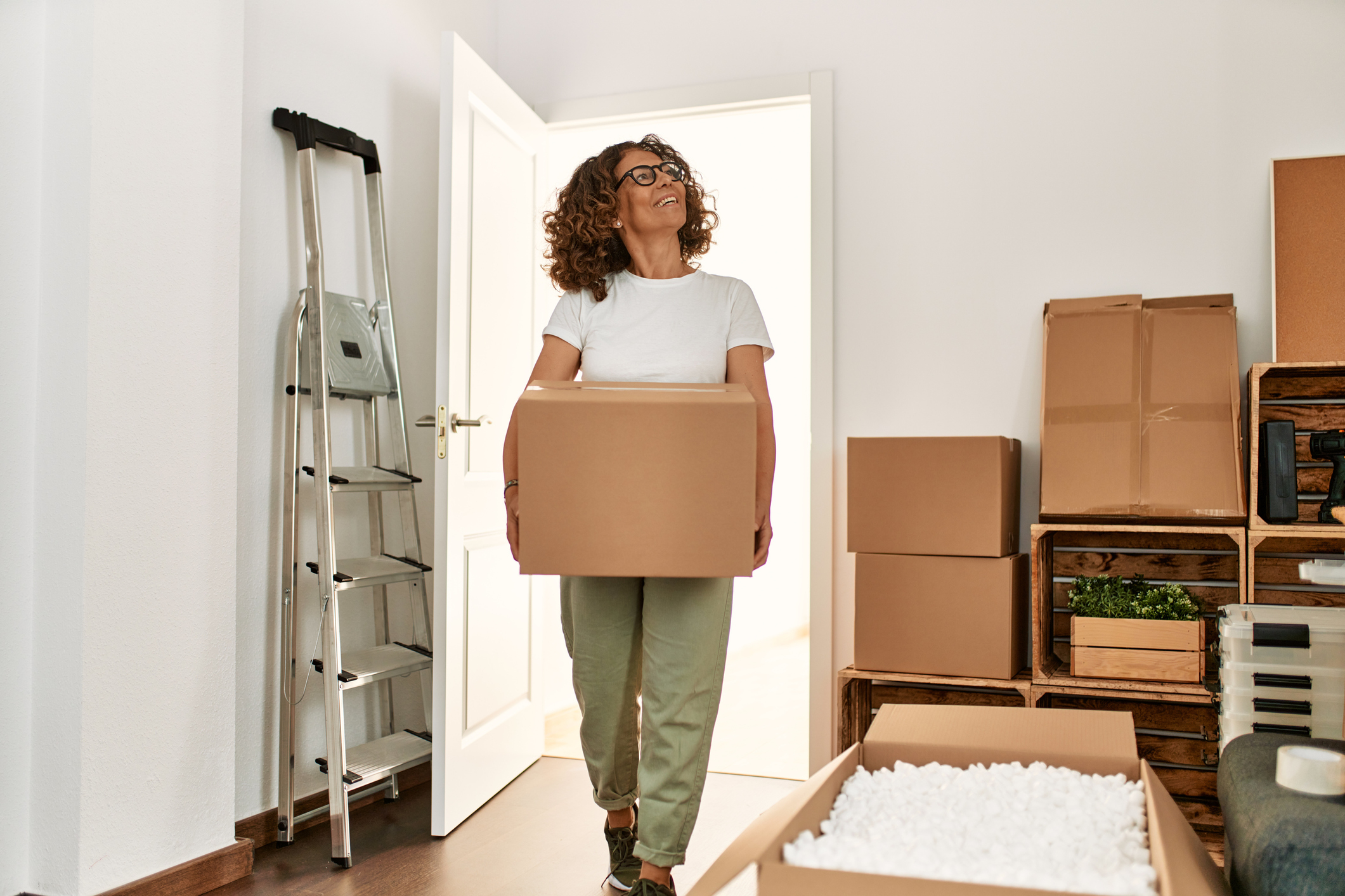 Woman with curly hair carries a box into a room filled with packing boxes and a ladder, smiling and looking upwards