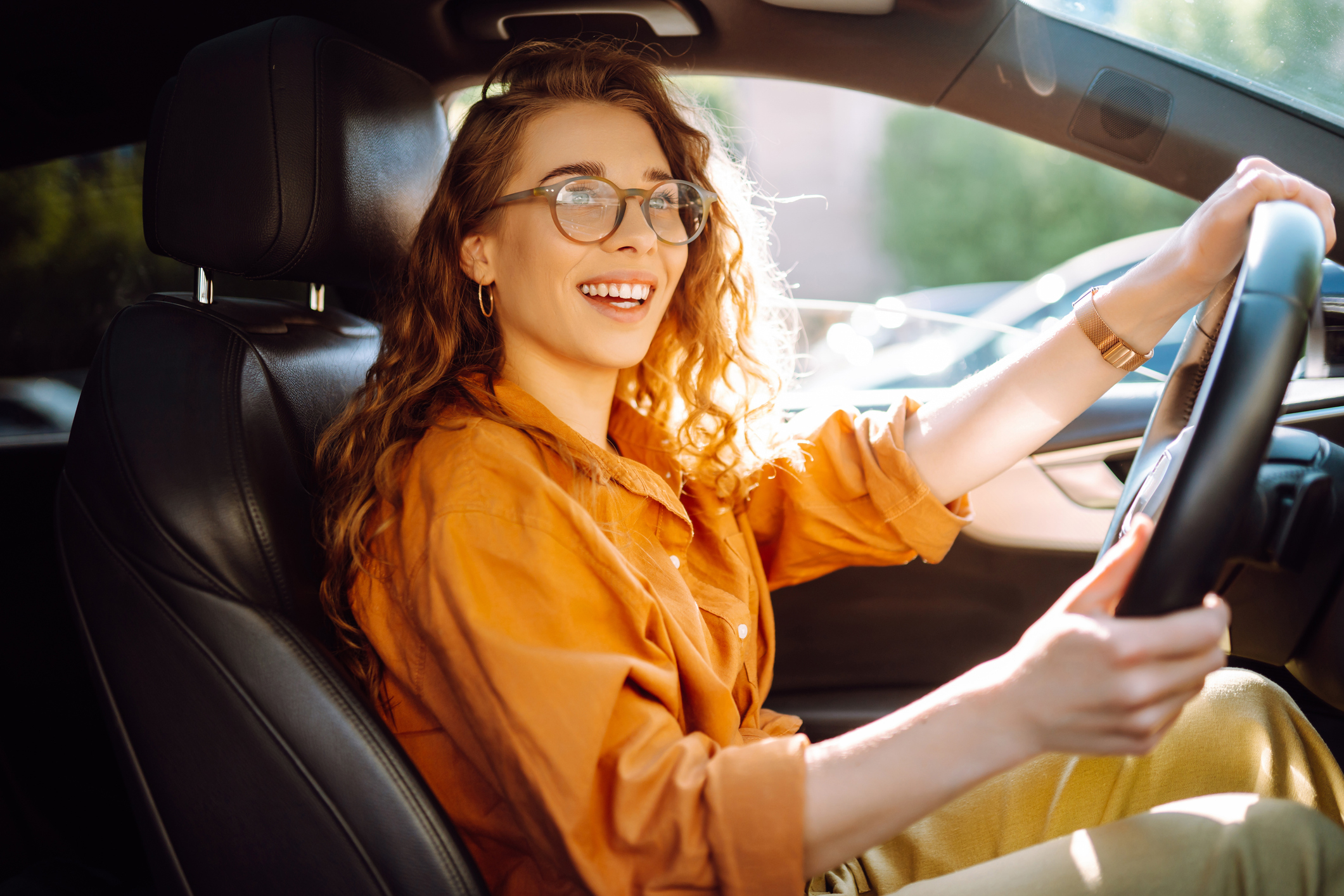 Person with long hair and glasses smiling while driving a car, wearing a casual button-up shirt