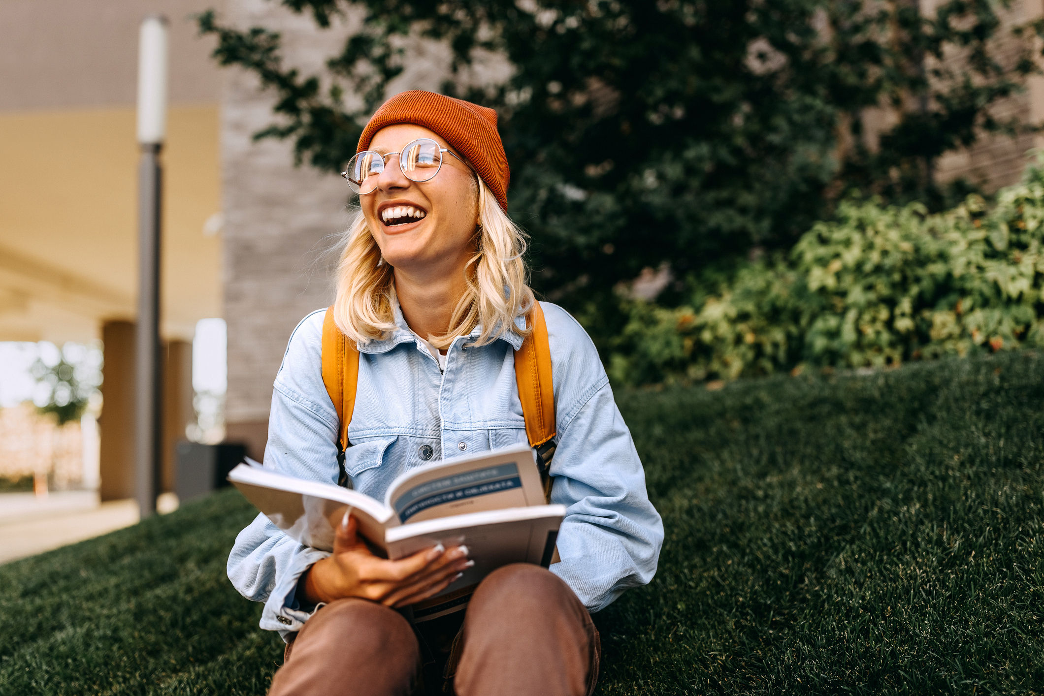 Person sitting on grass, smiling, wearing a beanie, glasses, denim jacket, and holding an open book. Trees and a building are in the background