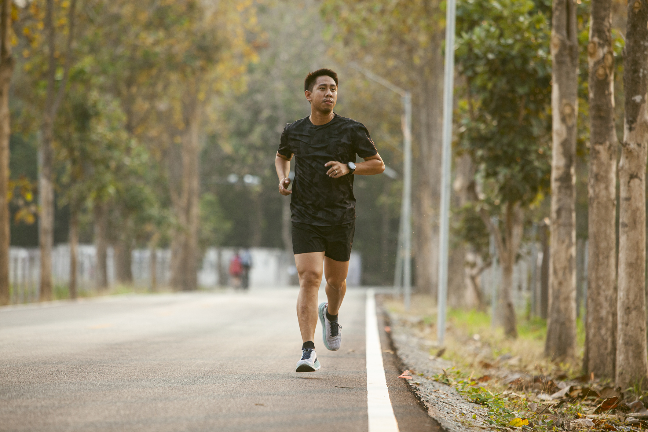 Man jogging on a tree-lined road, wearing a camo-patterned shirt and black shorts, focused on exercise